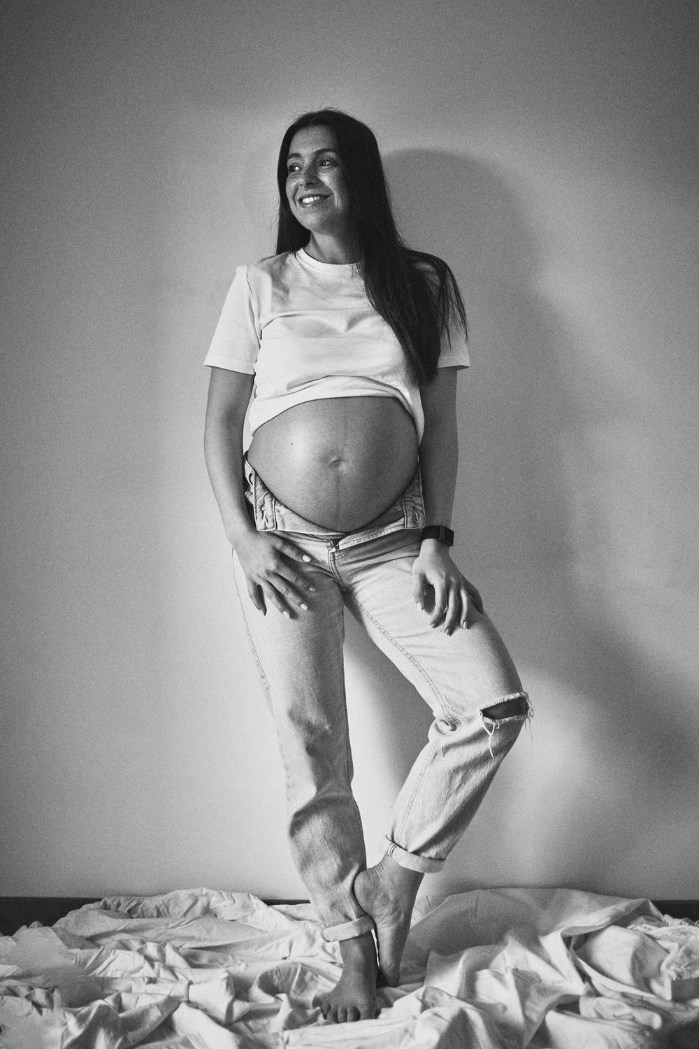 Black and white full body photo of smiling pregnant woman in white tee and ripped jeans.