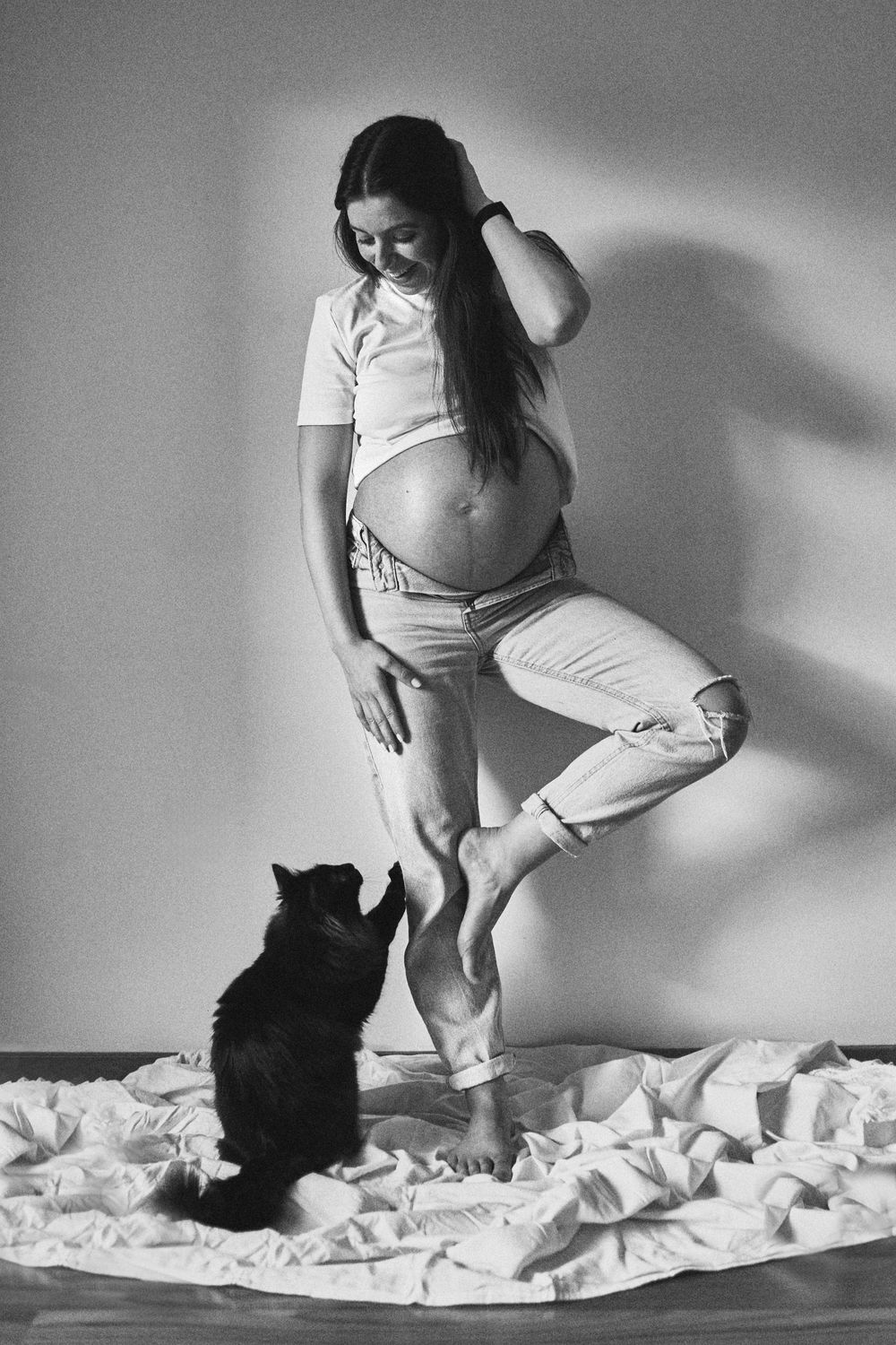 Black and white photo of pregnant woman in jeans with cat looking up at her baby bump.