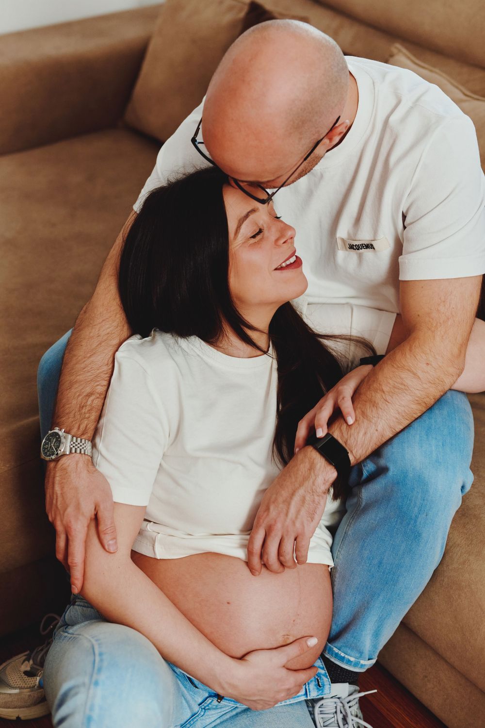 Tender moment as bald man kisses smiling pregnant woman while gently holding her belly.