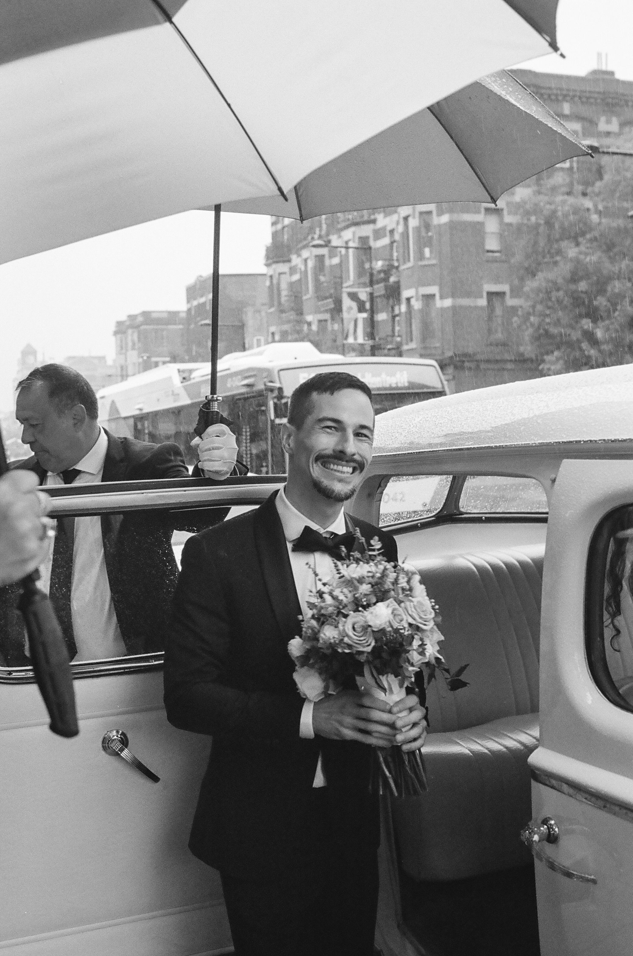Classic car with wedding party holding umbrellas in vintage black and white photograph.