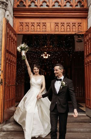 A couple exits a church with ornate wooden doors as confetti falls around them during their wedding celebration.