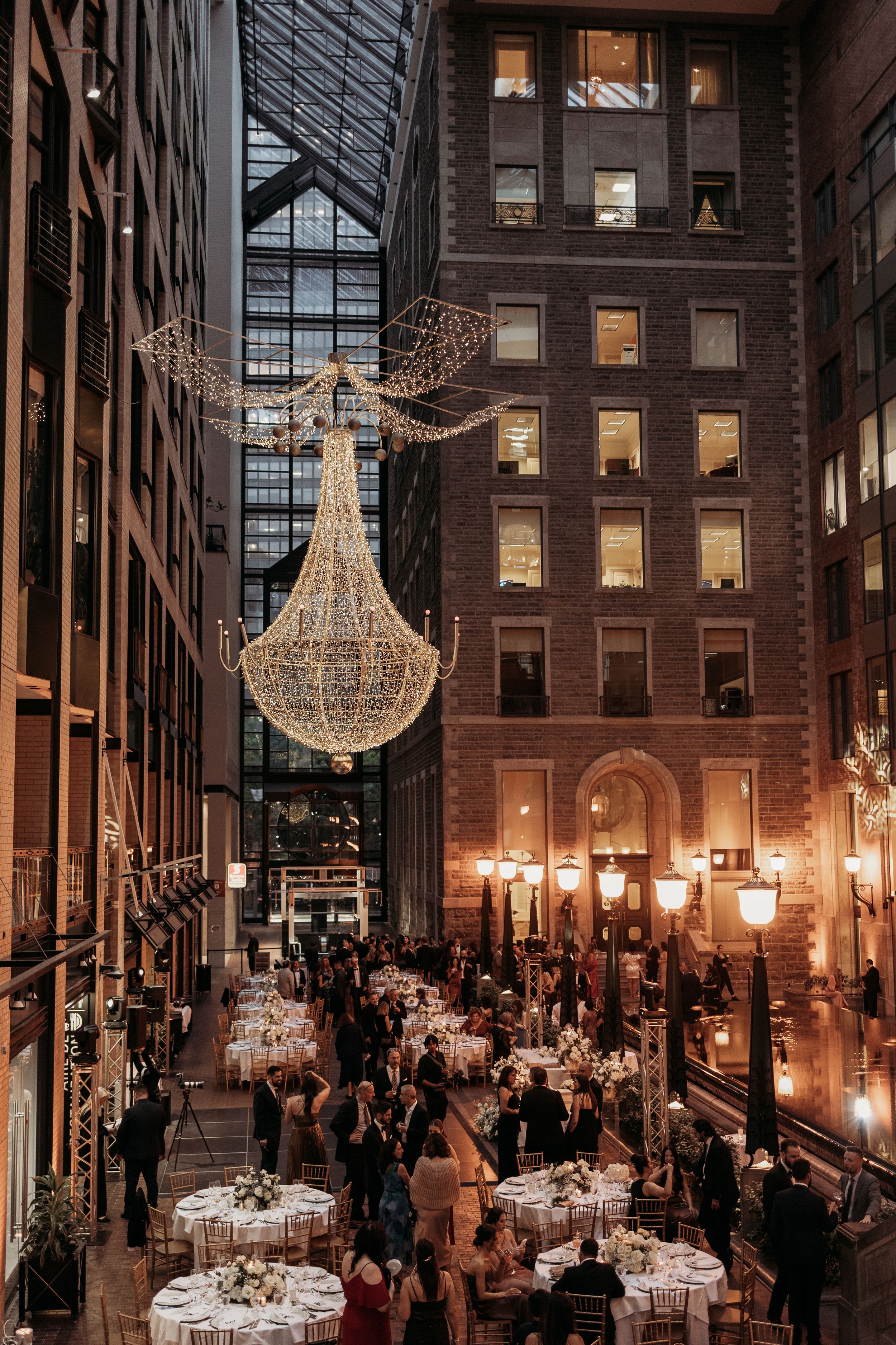 An elegant chandelier illuminates a formal indoor dining event with tables set beneath soaring ceilings in a historic building.