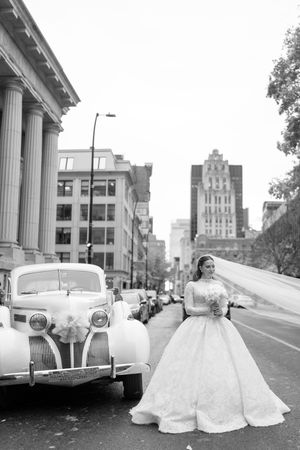 Vintage car parked in front of a classical building with white gown displayed in black and white photography.