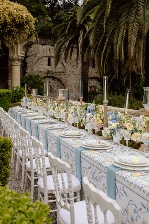 Elegant outdoor wedding table setting with blue and white decor, palm trees, and stone architecture in the background.