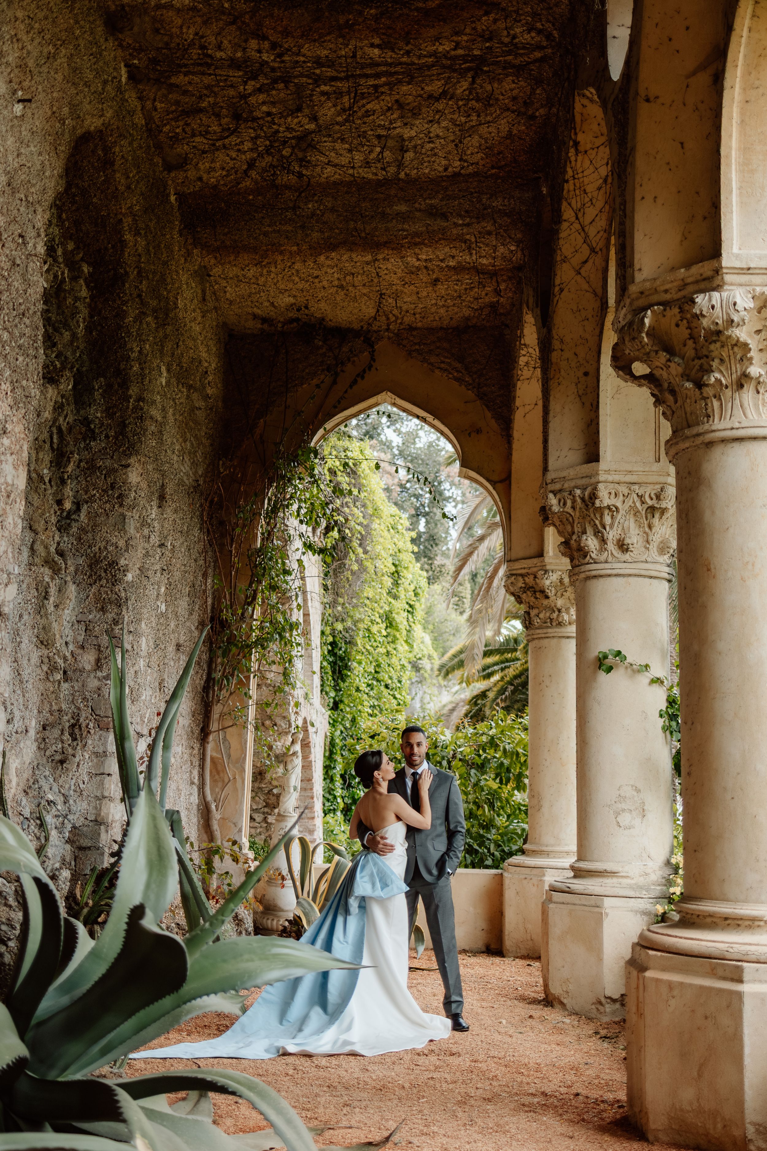 A romantic couple stands beneath ornate stone archways with trailing vines and agave plants in a Mediterranean garden setting.