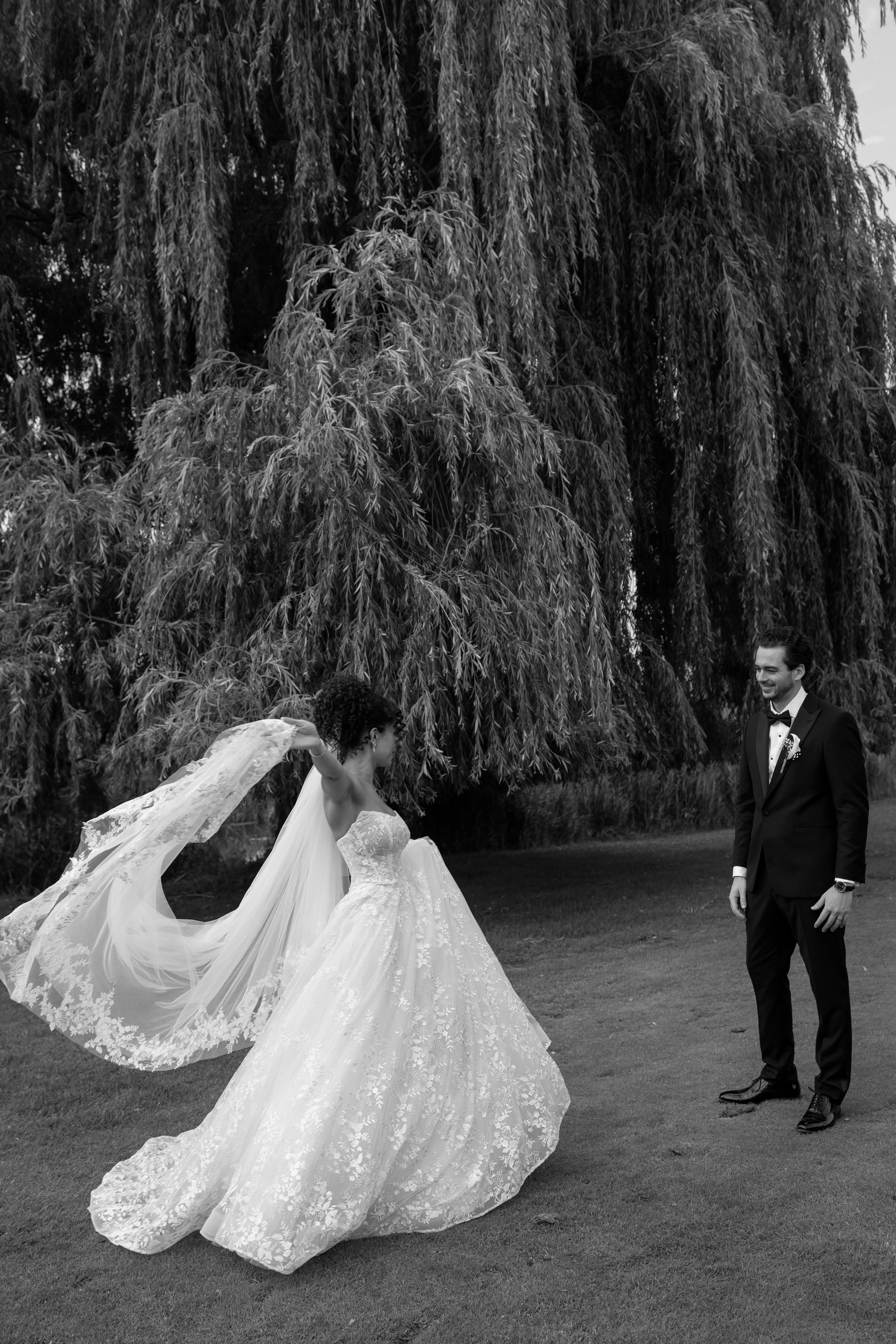 A black and white wedding photo series showing a flowing lace wedding dress train spread across grass under a weeping willow tree.