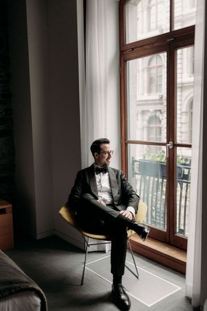 A person in formal attire sits thoughtfully by a large wooden window in a minimalist interior setting.
