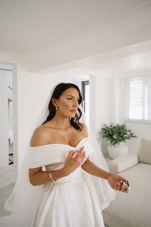 A bride in an off-shoulder white gown stands gracefully against a white wall in a sunlit room.