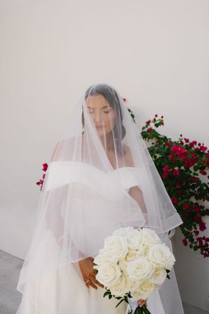 Elegant bride in flowing veil holds a stunning bouquet of white roses against a backdrop of red flowers.