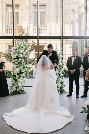 Wedding ceremony setup in bright venue with white floral arrangements and guests in formal attire.