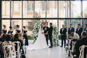 An elegant indoor wedding ceremony with white chairs and floral arch in a bright glass-walled venue.