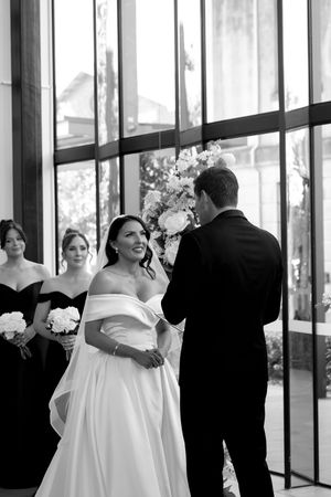 Wedding party in formal attire gathers in an elegant venue with floor-to-ceiling windows and white floral arrangements.