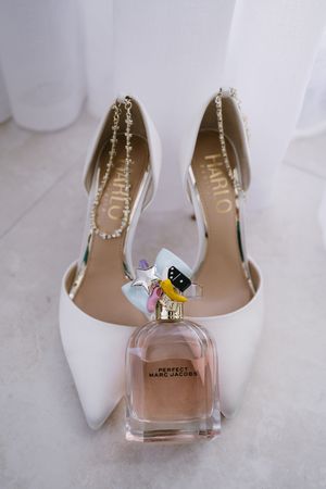 White wedding shoes displayed with a pink perfume bottle against a marble background.