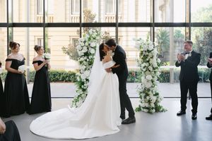 Wedding ceremony in progress at an elegant venue with floor-to-ceiling windows and floral decorations.