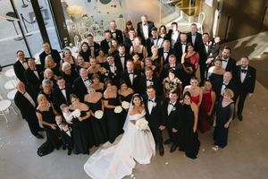Large wedding group photo with guests in formal black and burgundy attire gathered in an indoor venue.
