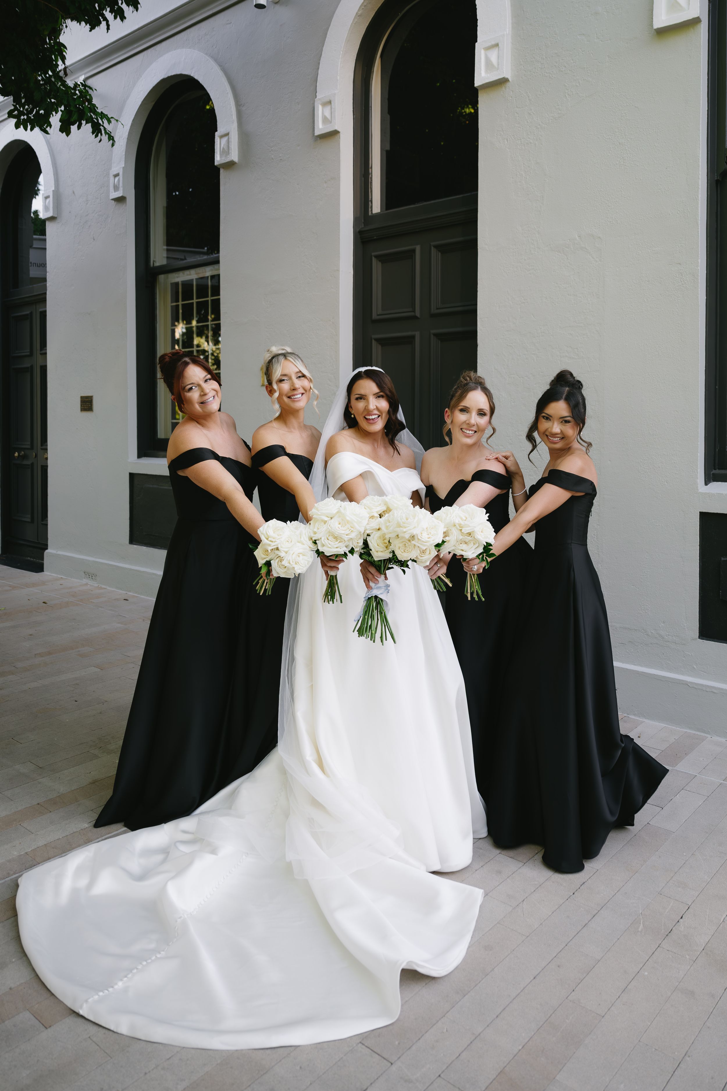 Wedding party poses together in elegant black bridesmaid dresses and white bridal gown holding white bouquets.