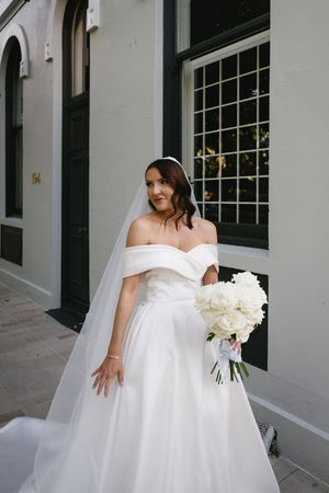 A bride in an elegant off-shoulder white wedding gown holds a bouquet of white flowers against a white building exterior.