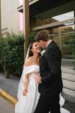 Romantic moment between newlyweds outside a modern building with glass windows and greenery.