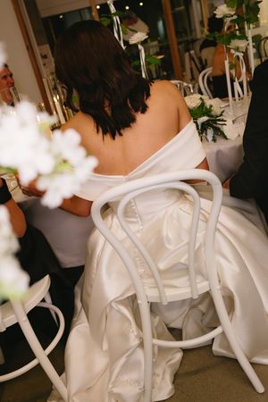 Back view of a bride in a white wedding dress seated at a table with floral decorations and white chairs.