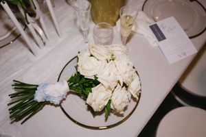 A classic white rose wedding bouquet rests on a formal place setting at a reception table.