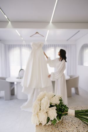 A white wedding dress hangs in a bright room with white roses and a robe-clad figure.