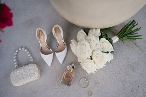 Wedding details flatlay with white heels, rings, roses and pearl accessories on a light gray background.