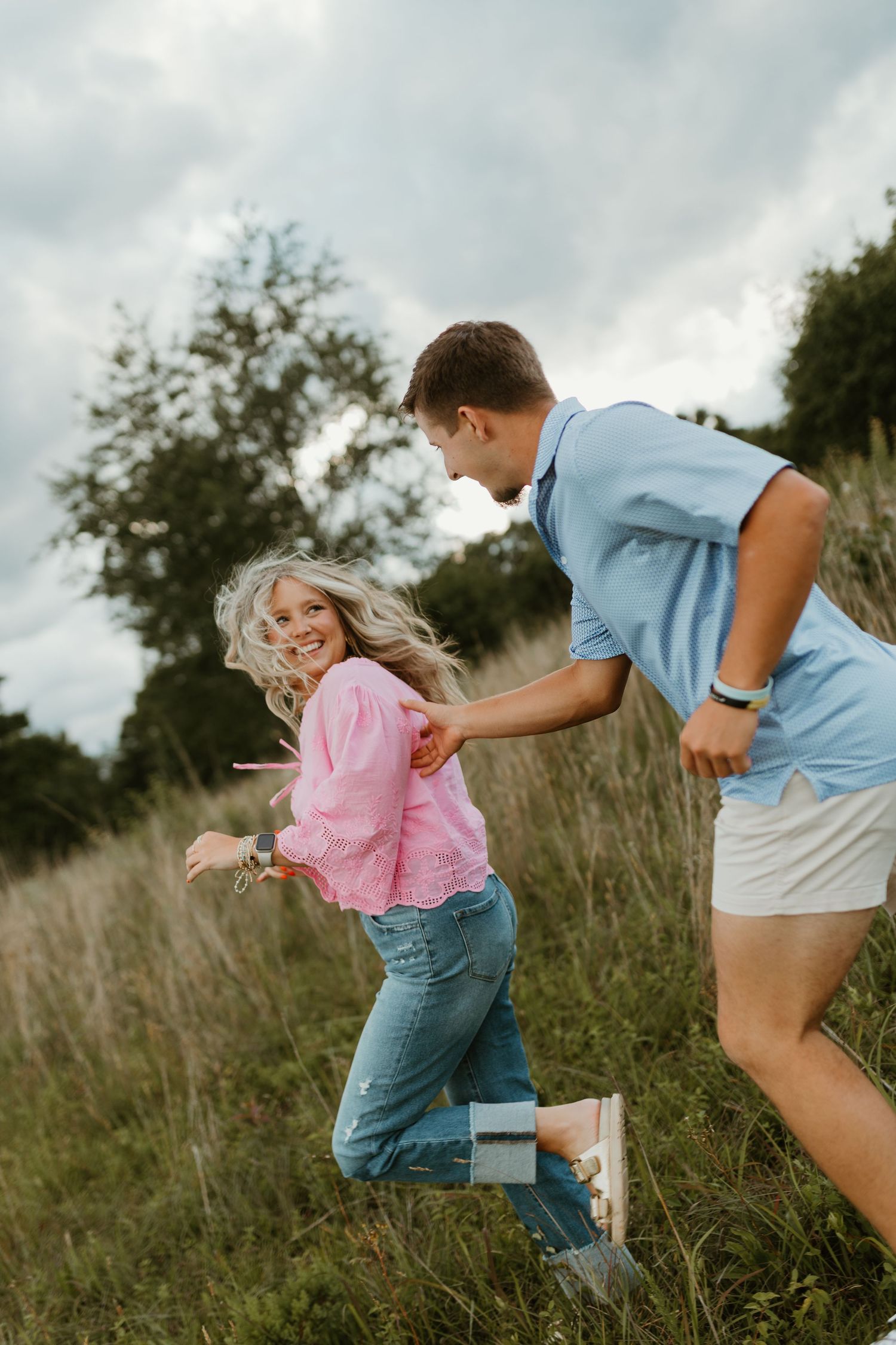 Playful couple running and chasing each other through a grassy field on a cloudy day.