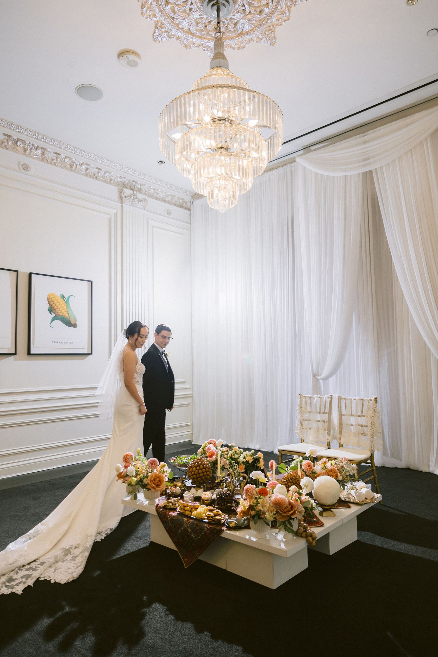 Elegant wedding venue with crystal chandeliers, white drapes, and floral centerpieces on tables against dark carpet.