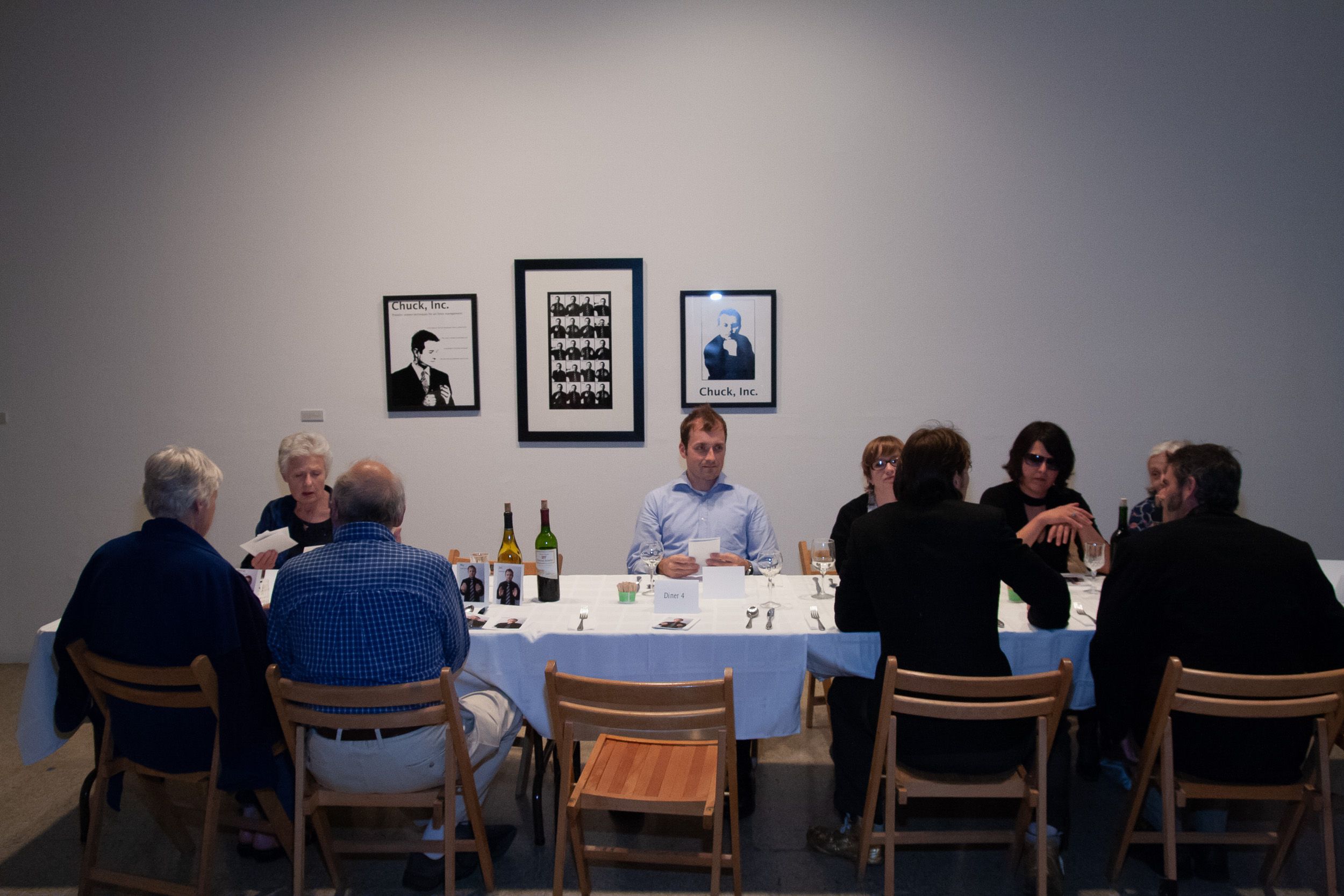 Gallery dining event with wooden chairs and white tablecloths under ambient lighting.