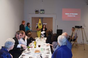 Corporate event showing attendees seated at tables with presentation screen visible in background.