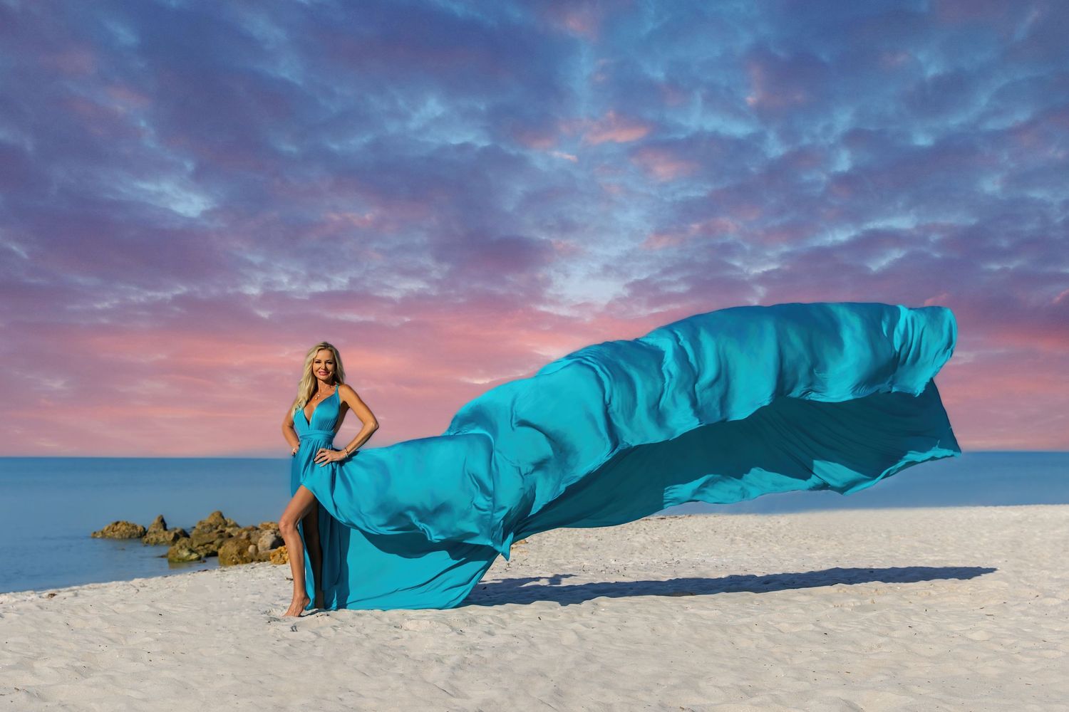 A flowing turquoise fabric creates a dramatic silhouette against a vibrant sunset sky on a white sandy beach.