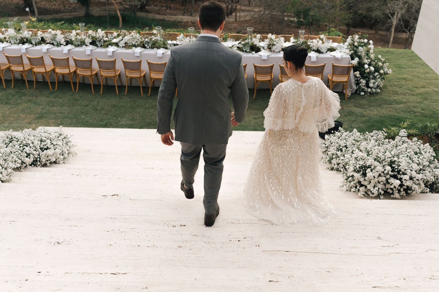 A couple walks together outdoors during a wedding celebration with white floral decorations and rustic seating.