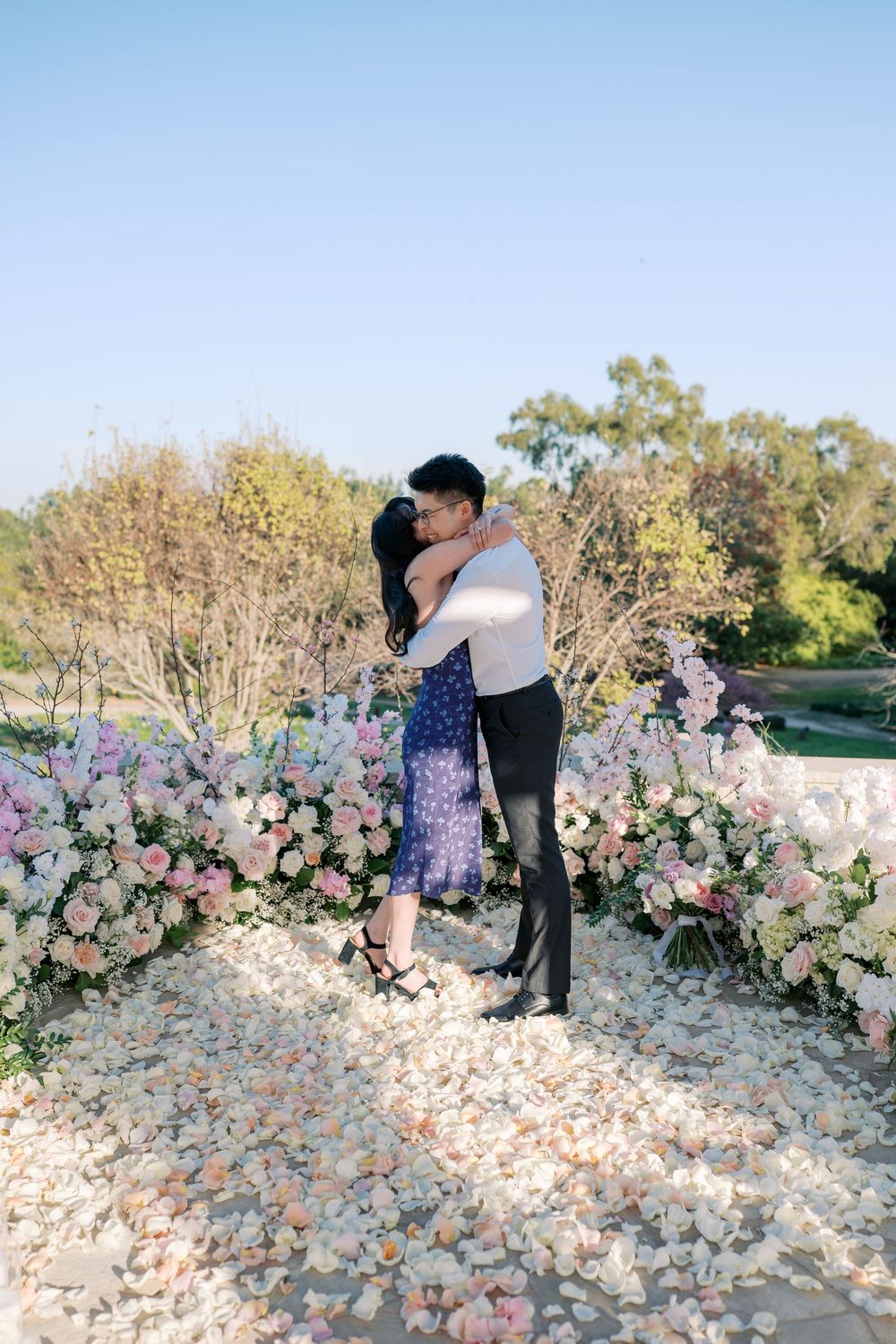 A romantic couple embraces surrounded by pink and white floral arrangements in an outdoor garden setting.