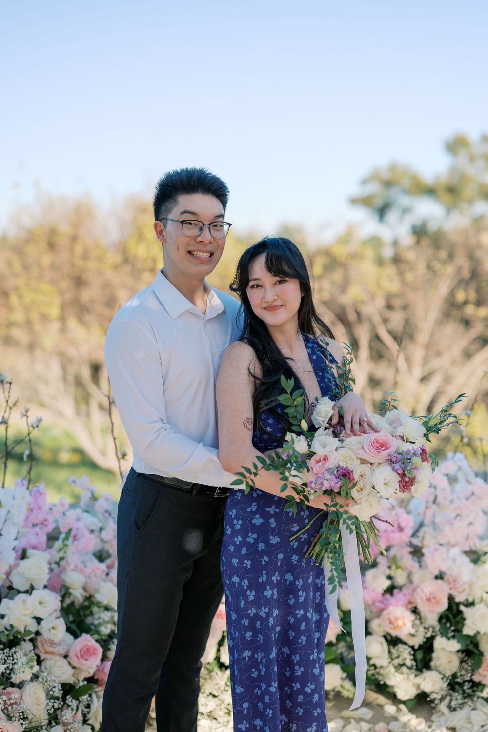 A couple poses together in an outdoor garden setting with pink floral arrangements and natural sunlight.
