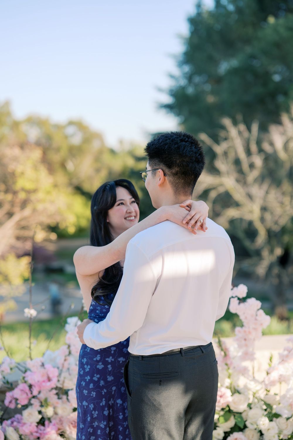 A couple shares a joyful moment together among pink floral decorations in a sunny garden.
