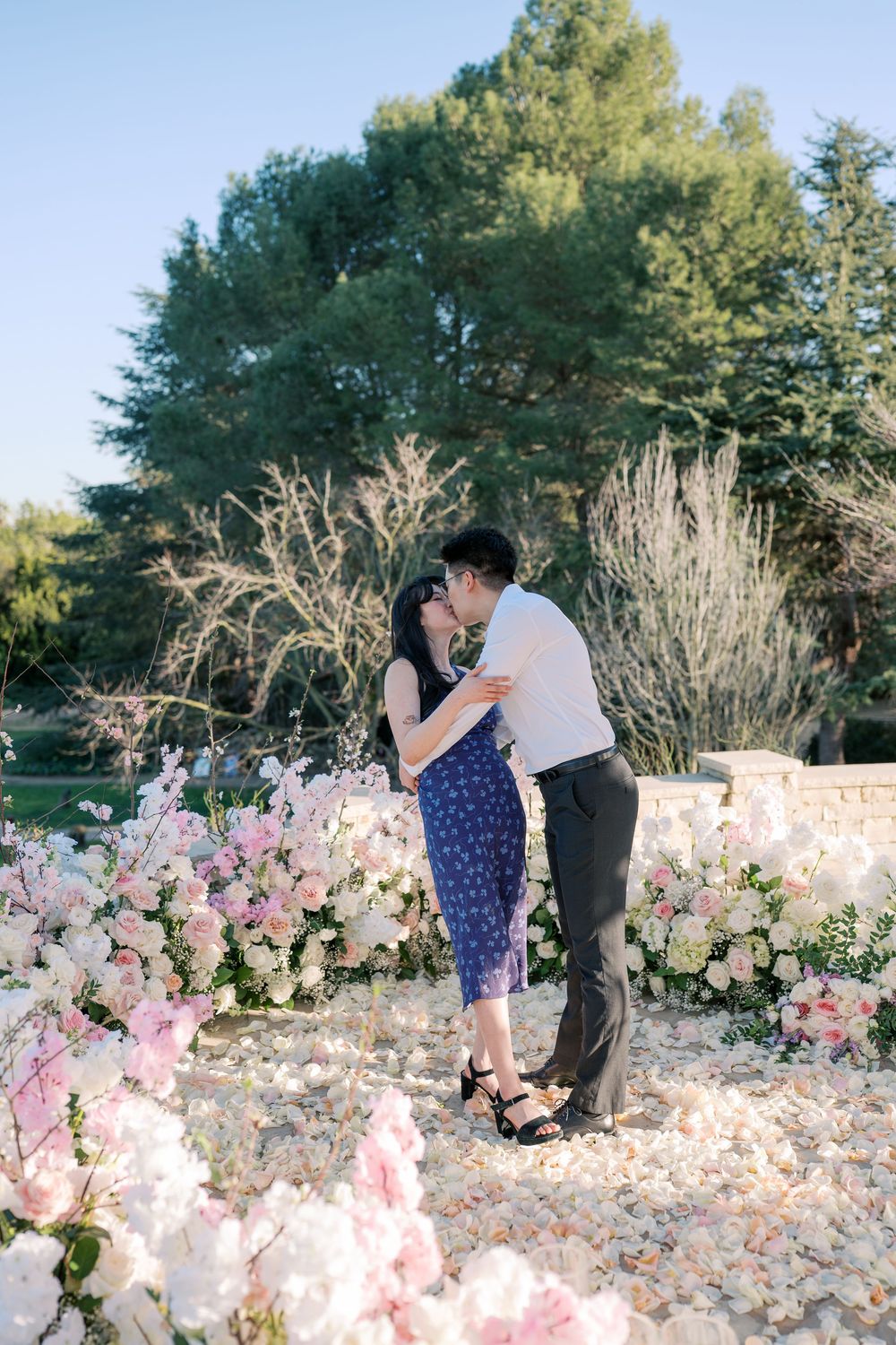 A couple shares an intimate moment surrounded by beautiful pink and white flower arrangements in a garden.