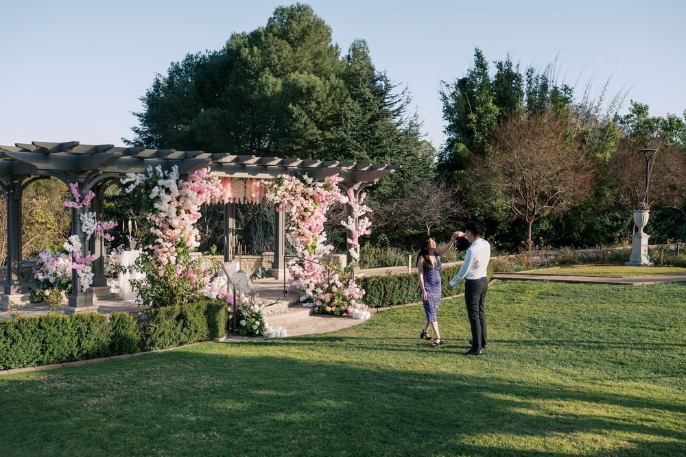 People gather in a lush garden setting with blooming cherry blossoms and green grass on a sunny spring day.