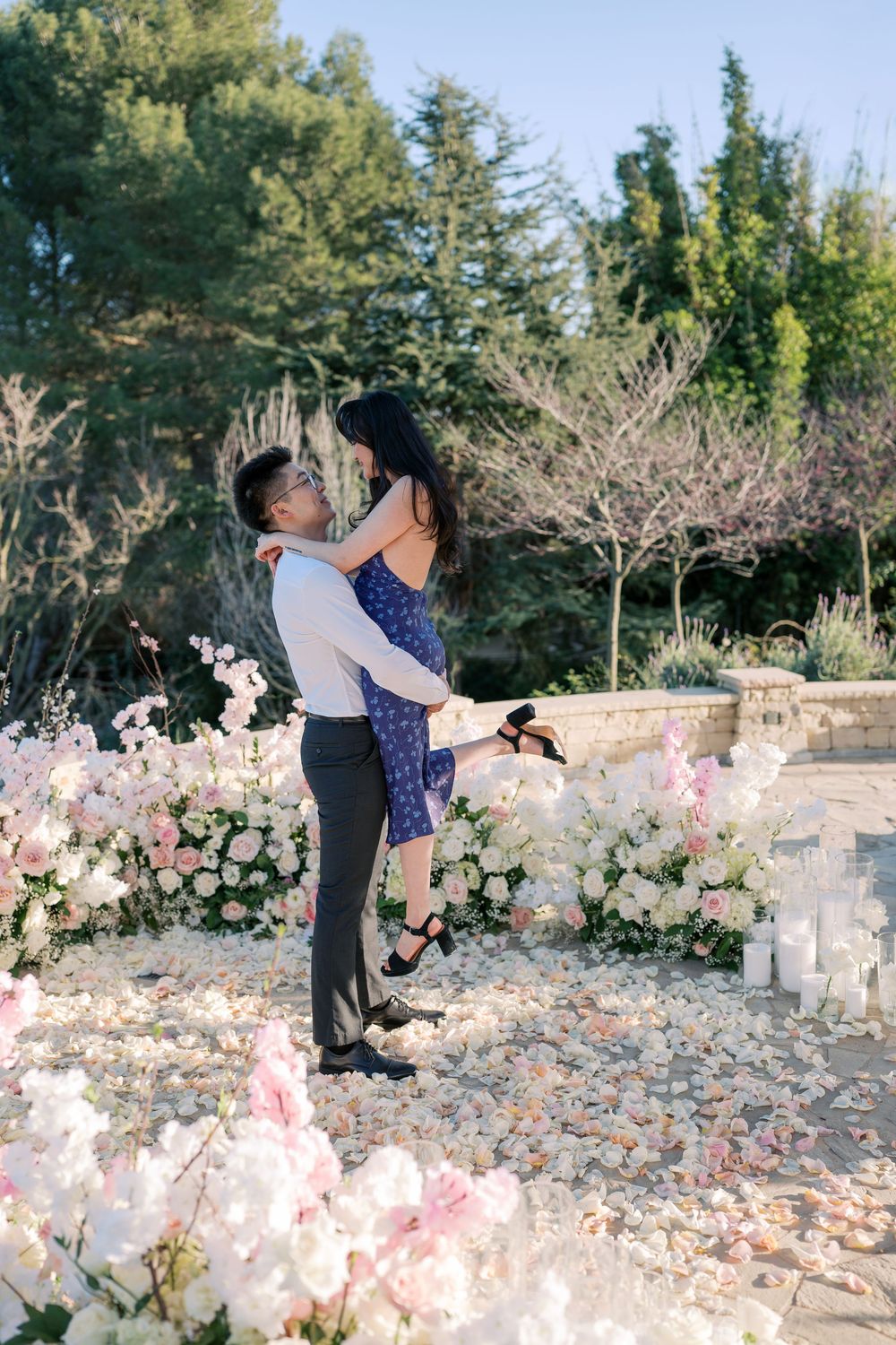 A romantic couple embraces among pink cherry blossoms in a garden setting with evergreen trees in the background.