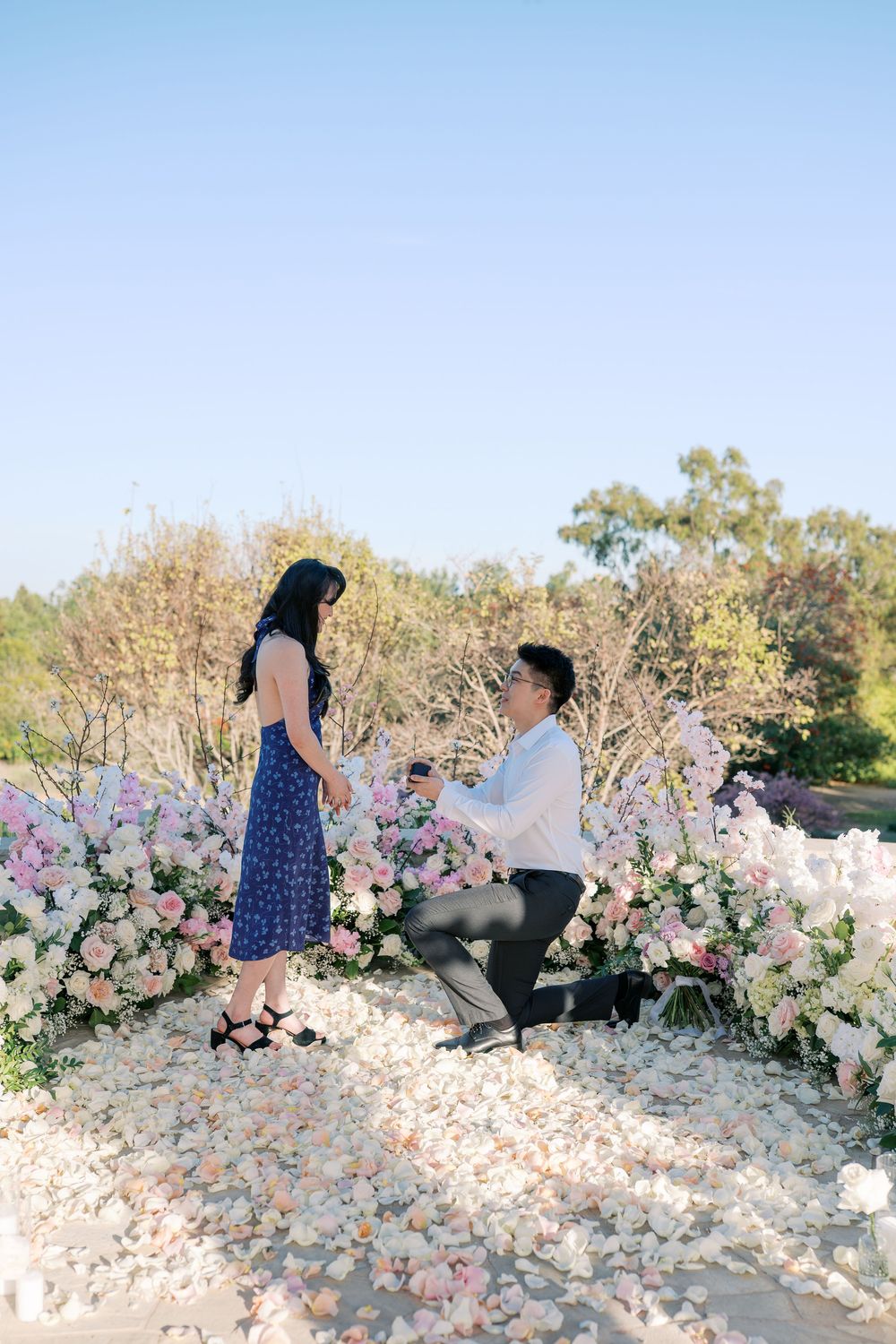 A romantic proposal scene unfolds in a garden setting decorated with pink and white floral arrangements.
