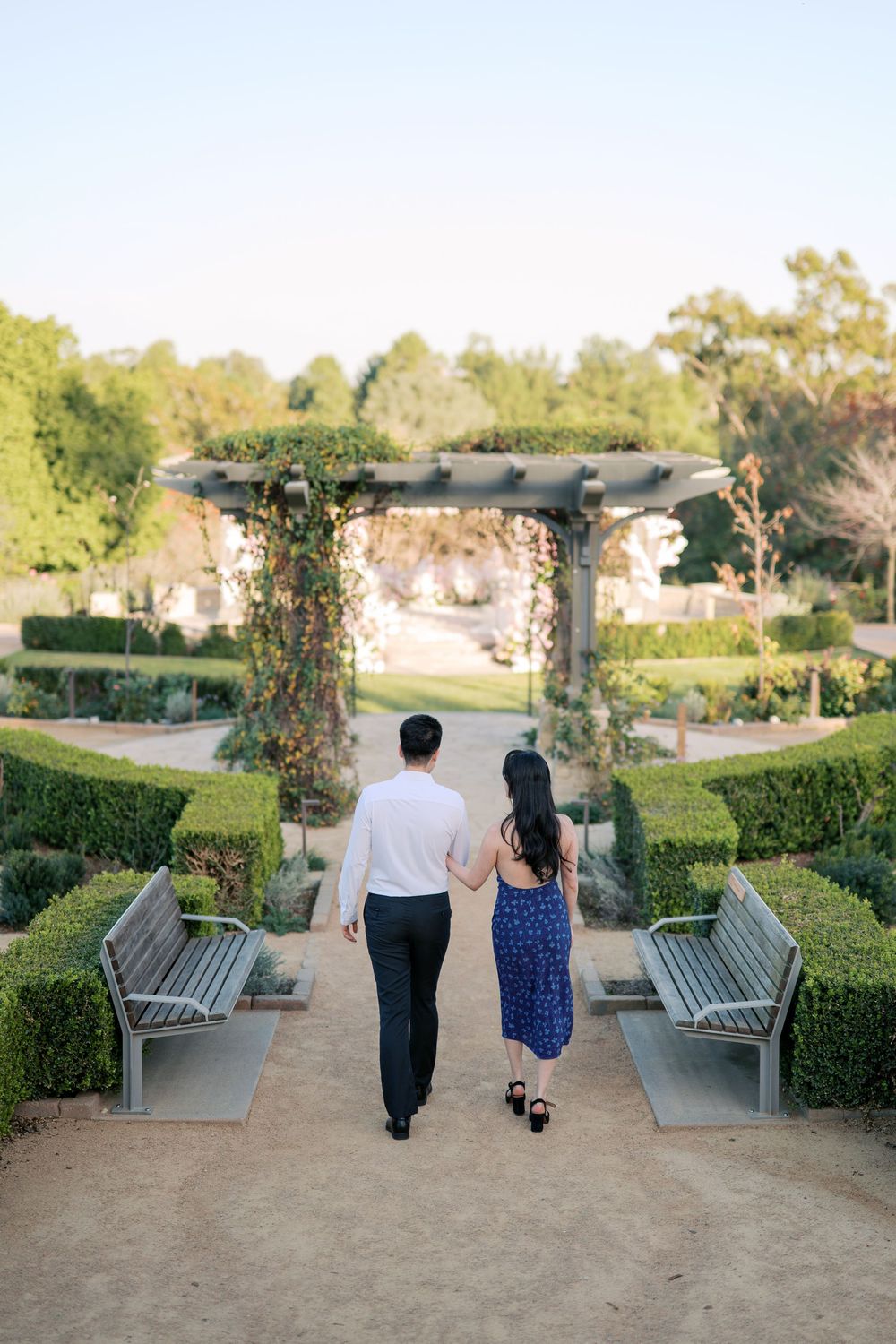 A couple walks away from the camera down a garden path with benches and hedges on a sunny afternoon.