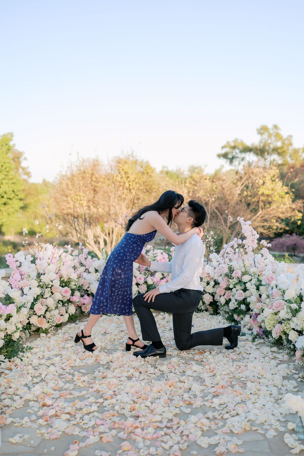 A romantic proposal scene with pink and white floral arrangements on the ground during sunset.