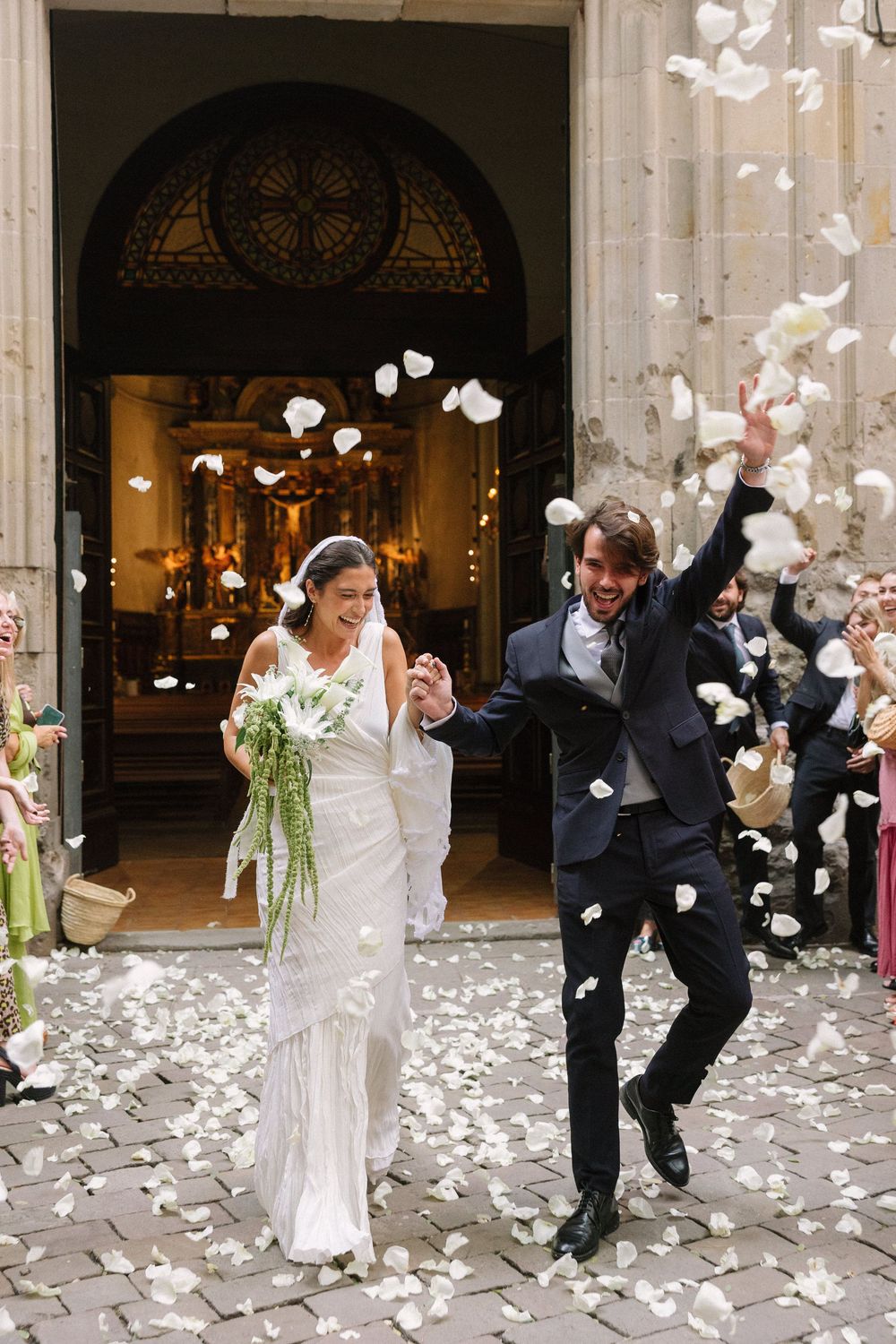 Wedding party members dodge and play in a shower of white flower petals during a celebratory recessional moment Gotic Barcelona.