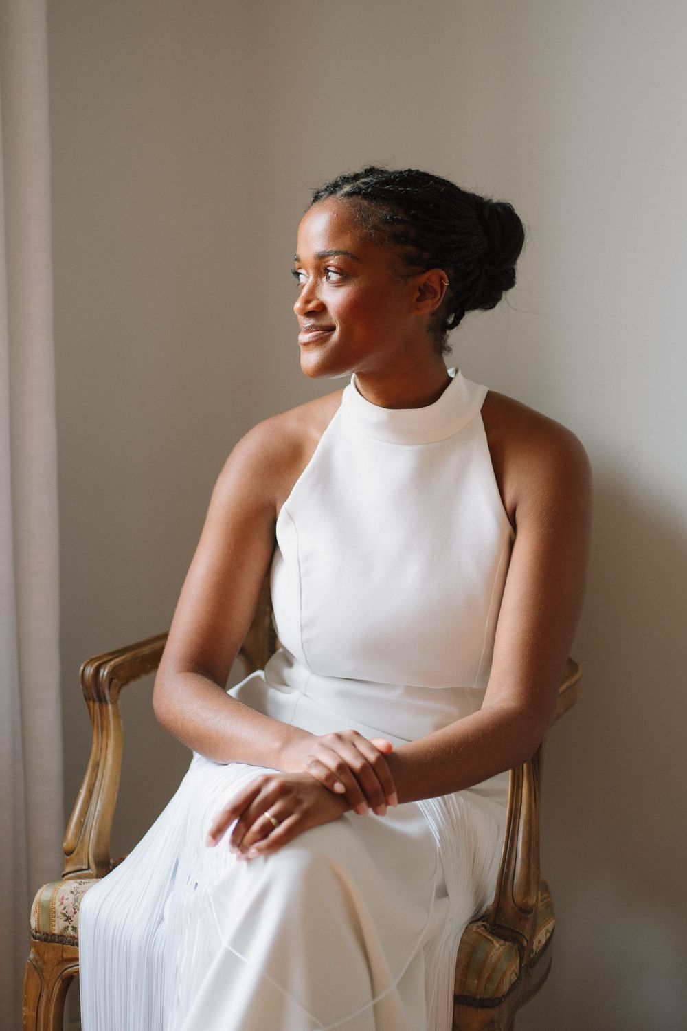 A woman in an elegant white halter dress smiles while sitting in an antique gold chair against a neutral wall.