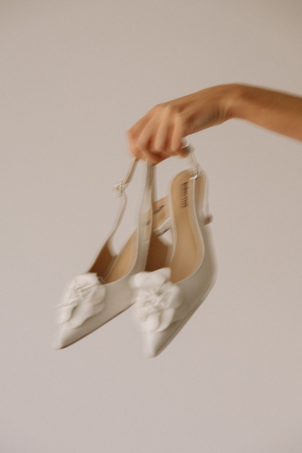 An elegant pair of white slingback shoes with bows being held up against a neutral background.