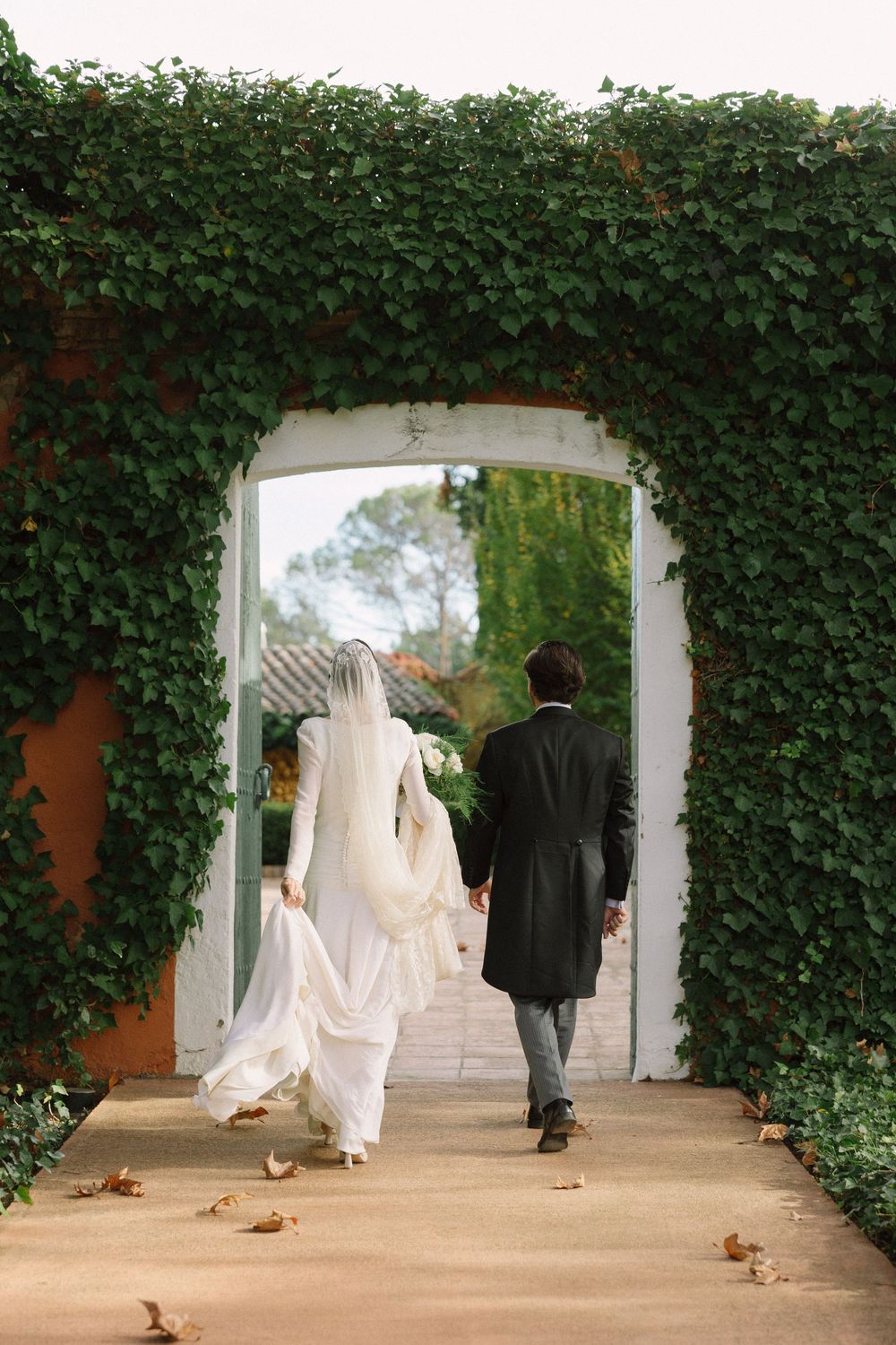Couple walking through an ivy-covered archway with autumn leaves scattered on the ground  Masia Rosas Sant Cugat.