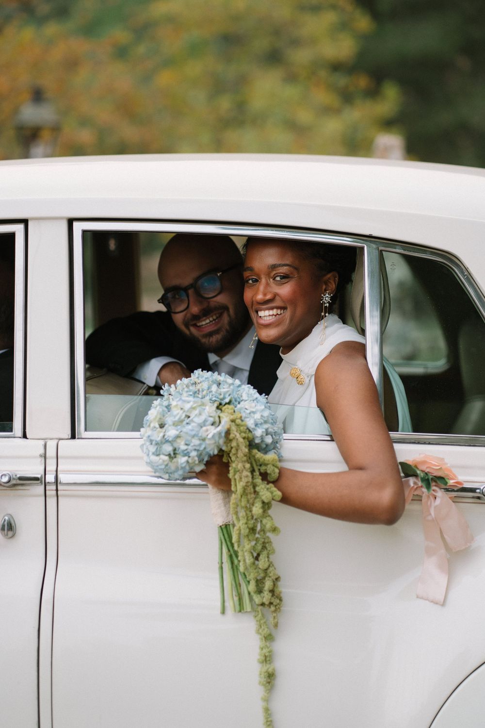Wedding party leaning out of vintage white car windows with blue hydrangea bouquets.