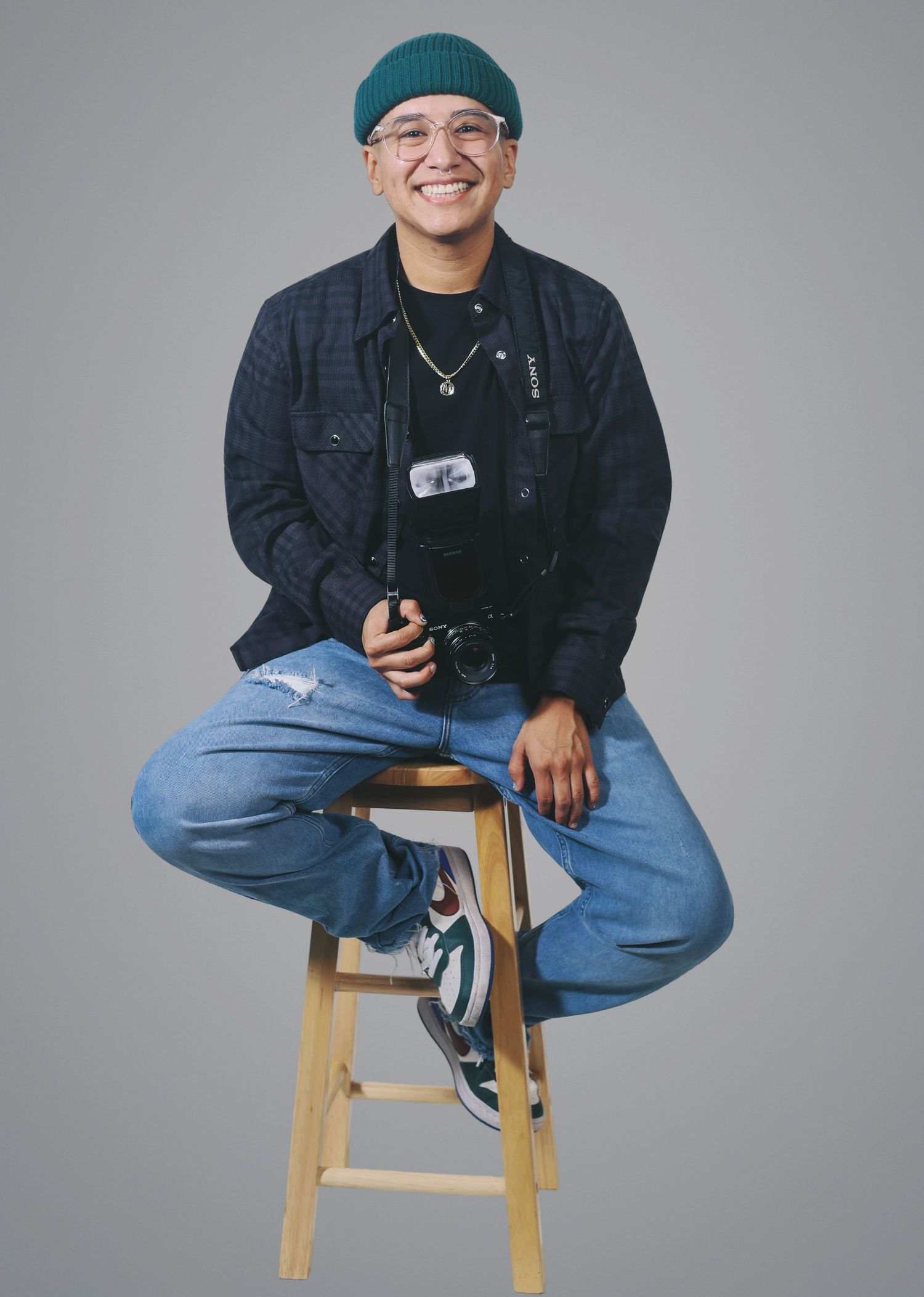 A photographer in a teal beanie and black jacket sits casually on a wooden stool against a white backdrop, holding a camera.