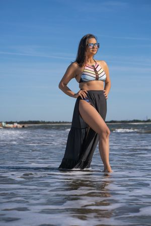 A person in a striped swimsuit and flowing black cover-up poses gracefully in shallow ocean waters on a sunny day.