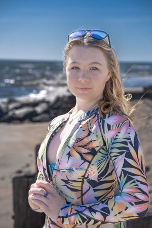 A person wearing a colorful tropical print jacket enjoys ice cream at a sunny beach with blue water in the background.
