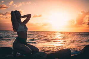 Silhouetted figure stands on beach during vibrant orange sunset over ocean waves.
